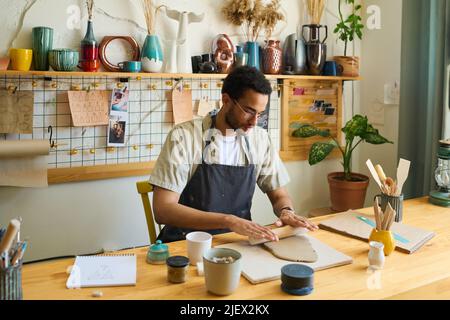 Junger multiethnischer Mann in Arbeitskleidung, der Ton abflacht, um neue handgefertigte Steingut-Artikel zu erstellen, während er am Arbeitsplatz sitzt Stockfoto