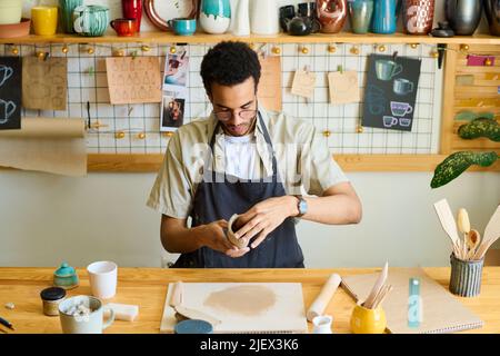 Junger kreativer schwarzer Mann in Arbeitskleidung, der neue Tonwaren zum Verkauf kreiert, während er in der Werkstatt am Tisch mit Steingut gegen das Regal sitzt Stockfoto