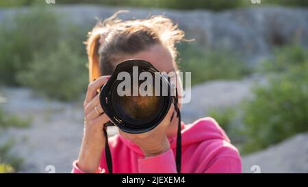 Junge Fotografin, die draußen steht und ein Foto mit professioneller Kamera direkt bei Ihnen fotografiert. Stockfoto