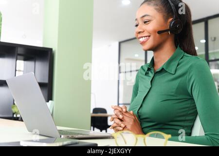 Junge, fröhliche afroamerikanische Frau, die im modernen Büro ein Headset für die Online-Kommunikation verwendet. Angenehme weibliche Betreiberin, die mit Kunden oder Kollegen auf der Ferne spricht Stockfoto