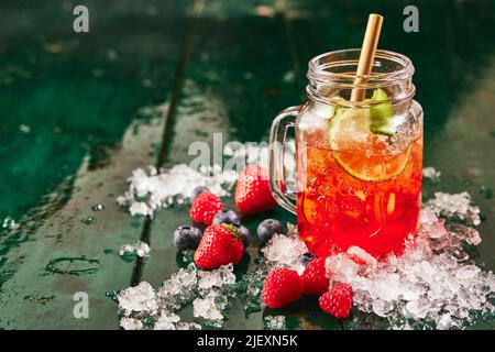 Glas gefüllt mit rotem Beerengetränk mit Limette serviert mit Stroh auf dem Tisch mit Eis und Erdbeeren und Heidelbeeren Stockfoto