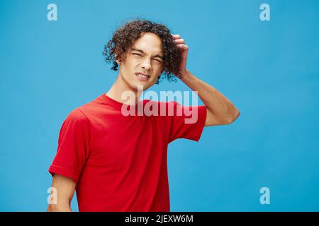 Nachdenklich nachdenklich unglücklich junge lockig gebräunt kaukasischen Kerl in grundlegenden roten T-Shirt posiert isoliert auf blauem Hintergrund. Lifestyle und Emotionen Stockfoto