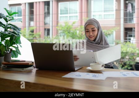 Die schöne und professionelle muslimische Buchhalterin mit Hijab sitzt an ihrem Schreibtisch und hält einen Papierkram-Bericht über ihre Konten. Stockfoto