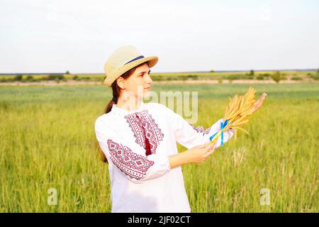 Unschärfe junge Frau in Vyshywanka und Hut hält Bouquet von reifen goldenen Ähren und Flagge auf der Wiese Natur Hintergrund. Flagge Ukraine. Fr. Stockfoto