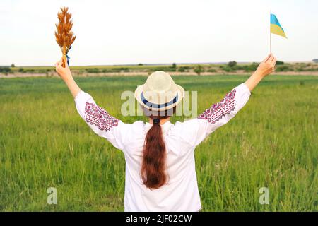 Unschärfe junge Frau in vyshywanka und Hut hält Bouquet von reifen goldenen Dornen von Weizen gebunden und Flagge auf der Wiese Natur Hintergrund. Flagge Ukrain Stockfoto