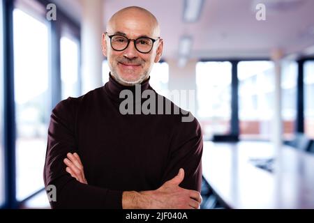 Legerer Geschäftsmann, der eine Brille trägt, während er mit gekreuzten Armen im Büro steht. Stockfoto