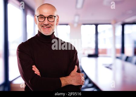 Legerer Geschäftsmann, der eine Brille trägt, während er mit gekreuzten Armen im Büro steht. Stockfoto