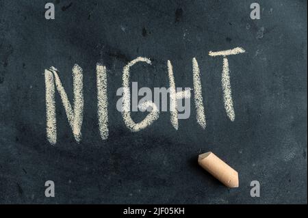NACHT. Der Name der Tageszeit ist in gelber Kreide auf einer schwarzen Tafel geschrieben. Der Text ist handschriftlich. Daneben ist ein Stück farbige Kreide. Stockfoto