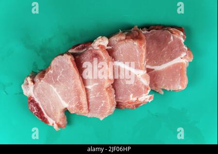 Rohes Schweinefleisch wird quer in portionierte Stücke geschnitten. Vier portionierte Stücke Schweineschulter. Rohes Fleisch liegt auf einer grünen, feuchten Oberfläche. Essen, Zutaten. Sele Stockfoto