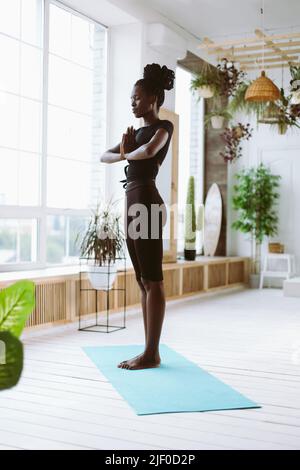 Vertikale freudige meditierende junge afroamerikanische Frau in schwarzer Sportkleidung Yoga praktizieren, halten Sie die Hände in namaste Mudra Stockfoto