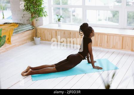 Rückansicht entspannende multirassische Frau in schwarzem Outfit, die Übungen macht, Yoga-Kobra-Pose am Fenster übt. Gesundheitswesen Stockfoto