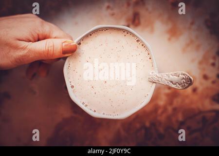 Eine Frau hält in ihrer Hand eine schöne graue Tasse mit warmem Kakao und Milch mit einem Teelöffel mit Mustern. Leckeres Getränk zum Frühstück. Stockfoto