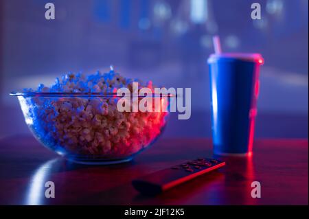 Popcorn in einer transparenten Schüssel, ein Getränk in einem Plastikglas und eine Fernbedienung auf einem Holztisch vor dem Fernsehbildschirm. Blaue Neonbeleuchtung. Res Stockfoto