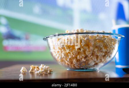 Stadion, Fußballplatz auf dem Großbildfernseher. Eine große Schüssel Popcorn auf einem Holztisch vor dem Fernseher. Fast Food, zu Hause beobachten Sportprogramme o Stockfoto