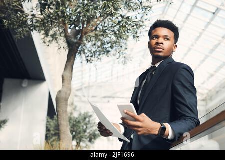 Selbstbewusster junger schwarzer Geschäftsmann in formeller Jacke, der sich in der modernen Lobby mit Bäumen auf Geländer stützt und ein profitables Geschäftsprojekt plant Stockfoto