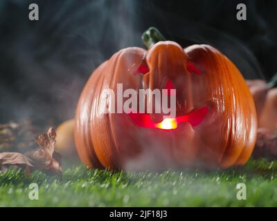 Orangefarbener Halloween-Kürbis mit einem unheimlichen Lächeln, von innen durch eine Kerze beleuchtet, verraucht auf einem grünen Rasen vor einem schwarzen, düsteren Himmel. Stockfoto