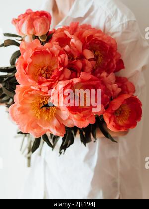 Junge Frau in weißen Kleidern hält ein großes Bouquet von korallenfarbenen Pfingstrosen. Vorderansicht Stockfoto