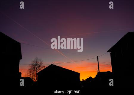 Gelber und orangefarbener Sonnenuntergang mit Gebäuden, Dächern und Bäumen, die sich gegen die Wolken und den blauen Himmel schilten Stockfoto