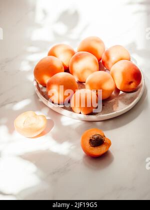 Reife Bio-Aprikosen auf weißem Teller im Sonnenlicht und den Schatten der Blätter des Baumes Stockfoto