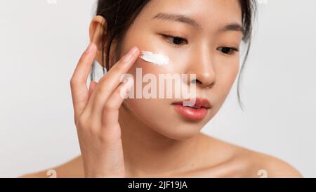 Nahaufnahme einer asiatischen Dame, die Anti-Aging- oder feuchtigkeitsspendende Gesichtscreme auf die Wange anwendet und auf weißem Studiohintergrund steht Stockfoto