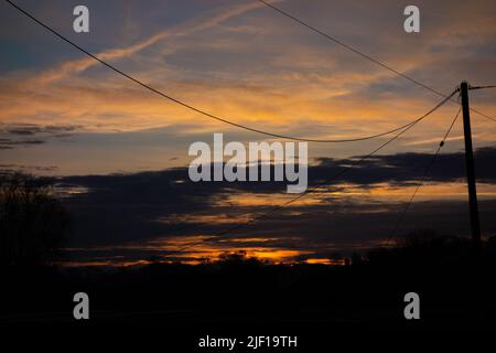 Gelber und orangefarbener Sonnenuntergang mit Stromleitungen und Bäumen, die sich gegen die Wolken und den blauen Himmel schilten Stockfoto