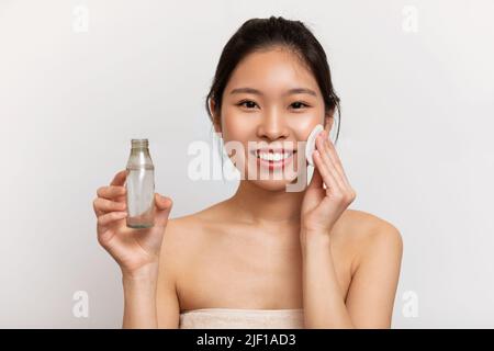 Fröhliche koreanische Dame mit Wattepad und Flasche mit Gesichtsreinigungsgel, die auf weißem Studiohintergrund steht Stockfoto