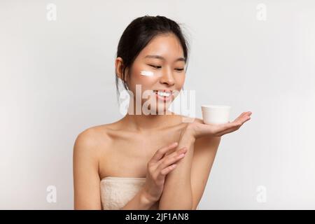 Glad asiatische Dame mit perfekter Haut Anwendung feuchtigkeitsspendende Creme auf das Gesicht und halten Glas mit leerem Raum, weißen Hintergrund Stockfoto