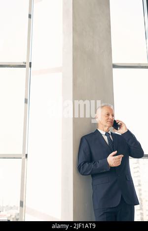 Inhalt ein gutaussehender, reifer Geschäftsmann in formellem Anzug, der sich in der Lobby auf eine Säule stützt, während er mit dem Handy spricht Stockfoto