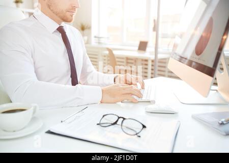 Beschnittenes Bild eines konzentrierten jungen Geschäftsmannes in formellem Hemd, der am Schreibtisch sitzt und am Computer Diagramme erstellt Stockfoto