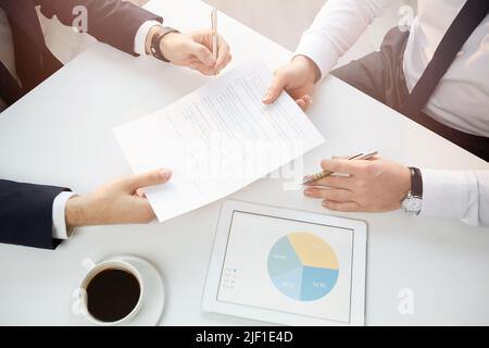 Oben ein unerkennbarer Business Manager, der mit einer Kaffeetasse und einem Tablet am Tisch sitzt und einen Vertrag mit einem Partnerschaftsvertreter bei busi unterzeichnet Stockfoto