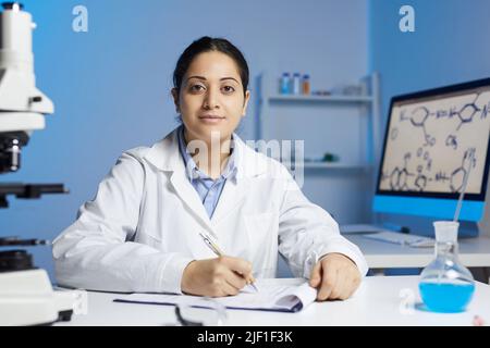 Content-Portrait junge indische Laborexpertin in weißem Mantel sitzt am Tisch im Büro und macht Aufzeichnungen über Experiment Stockfoto