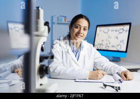 Porträt einer lächelnden jungen Inderin im Laborkittel, die am Schreibtisch sitzt und Notizen in der Zwischenablage macht, während sie im wissenschaftlichen Labor arbeitet Stockfoto