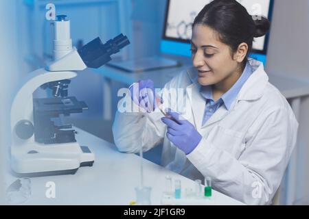 Positive junge indische Forscherin im Labormantel, die mit dem Mikroskop am Schreibtisch sitzt und mit Chemikalien arbeitet Stockfoto