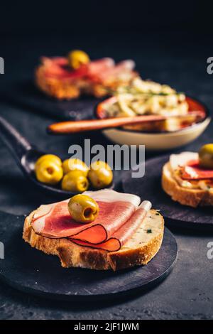 Geröstetes Brot oder offenes Sandwich mit Knoblauchbutter-Aufstrich und serano-Schinken und Oliven auf dunklem Hintergrund Stockfoto