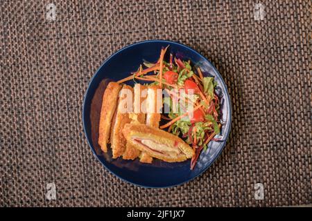 Köstlicher Salat aus grünem und rotem Gemüse, begleitet von panierten Hähnchen, gefüllt mit Schinken und Käse auf einem dunklen Teller Stockfoto