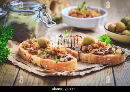 Frische hausgemachte Hähnchenleber-Pastete auf Ciabatta-Brot mit gerösteten Zwiebeln und Oliven auf einem Holztisch Stockfoto