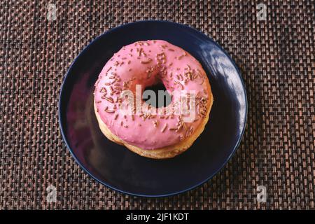 Köstliches rundes Brot mit rosa Glasur und bunten Streuseln auf einem dunklen Teller, bereit zum Frühstück Stockfoto