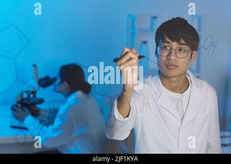 Konzentrierter, intelligenter asiatischer Chemiker in Laborkittel und -Gläsern, der an glasigen Brettschichten steht und ein Zeichenschema der molekularen Einheit zeichnet Stockfoto