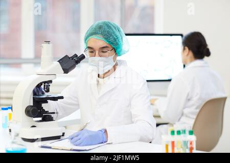 Seriöse junge asiatische Laborforscherin in steriler Arbeitskleidung, die mit dem Mikroskop am Schreibtisch sitzt und Aufzeichnungen über die Probe führt Stockfoto