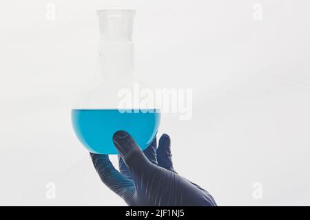 Hand eines nicht erkennbaren chemischen Forschers in einem Gummihandschuh, der ein Becherglas aus blauer Substanz vor weißem Hintergrund hält Stockfoto
