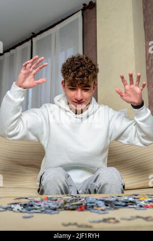 Ein junger kaukasischer Mann mit lockigen Haaren sitzt zu Hause auf dem Sofa und seine Arme sind angehoben, um gegen die Schwierigkeit zu protestieren, ein Puzzle von 1000 zusammenzustellen Stockfoto