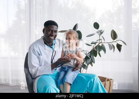 Ein afroamerikanischer Arzt kommuniziert mit einem kleinen Mädchen, das auf dem Schoß des Arztes sitzt Stockfoto