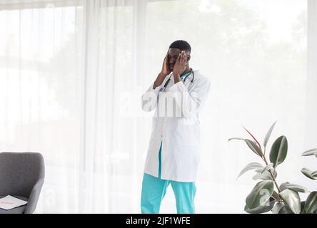 Ein afroamerikanischer Arzt, der von harter Arbeit müde ist, steht neben dem Fenster. Stockfoto