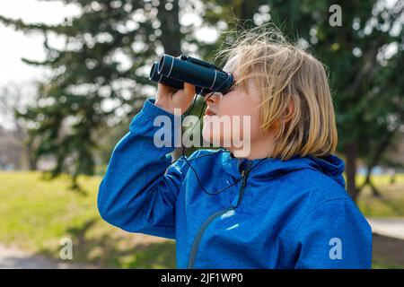 Kind schaut durch ein Fernglas im Park. Porträt eines kleinen Jungen, der die Tierwelt erkundet. Vogelbeobachtung, Wandern und Abenteuerkonzept. Stockfoto