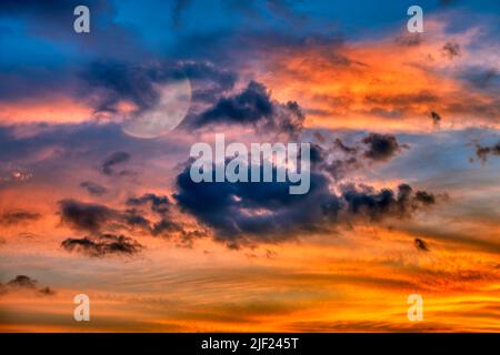 Der Mond steigt inmitten Eines farbenprächtigen, wolkigen Sonnenuntergangshimmels auf Stockfoto