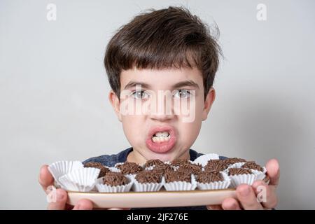 9-jähriges Kind hält ein Tablett mit mehreren brasilianischen Fudge-Kugeln und knirschen seine Zähne. Stockfoto