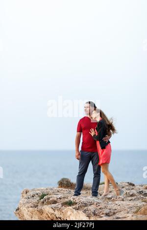 Porträt eines hübschen Paares mittleren Alters, das neben den Copyspace schaut und sich umarmt, während es am Rand der Felsklippe am Meer steht. Stockfoto