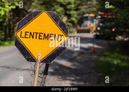 Nahaufnahme selektive Fokusansicht eines gelben Warnzeichens auf der Straße mit Wortlentement, Französisch für langsam. Verschwommener Bagger und LKW im Hintergrund gesehen. Stockfoto