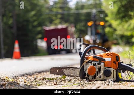 Nahaufnahme einer Motorsäge am Rande einer Hauptstraße mit selektivem Fokus. Verschwommene Bremsleuchten eines Baggers und eines Muldenkipper sind im Hintergrund zu sehen. Stockfoto