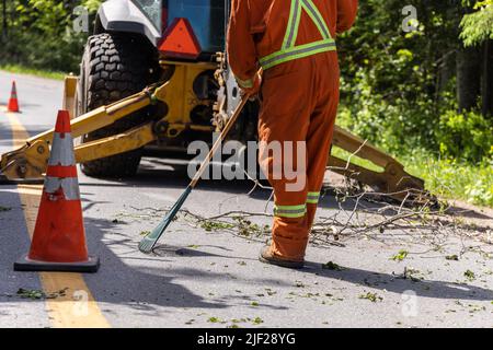 Nahaufnahme, während ein Arbeiter in gut sichtbarer Kleidung mit einem Rechen Baumzweige und Blätter von der Straße mit Traktor im Hintergrund entfernt. Stockfoto